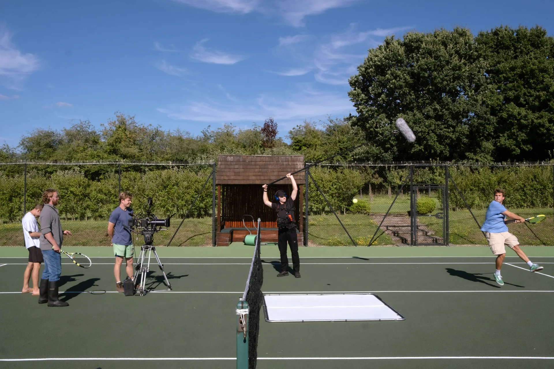 Joey Bader playing tennis while the camera and sound crew film across the net