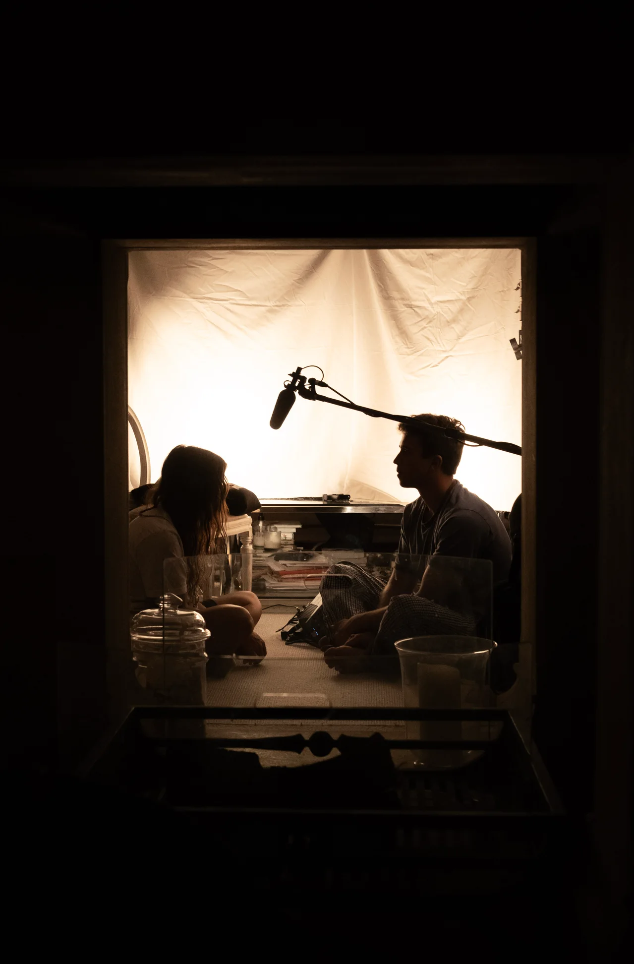 Two actors seated opposite each other beneath a boom, silhouetted against a large diffusion panel